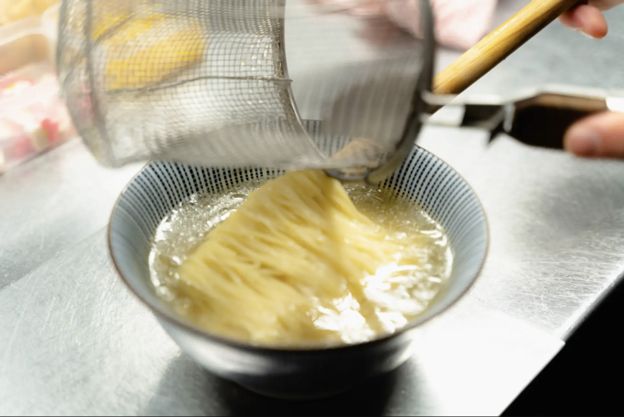 厨房で茹で上がった特注のストレート細麺を平ざるで丁寧に丼のスープへ盛り付ける様子